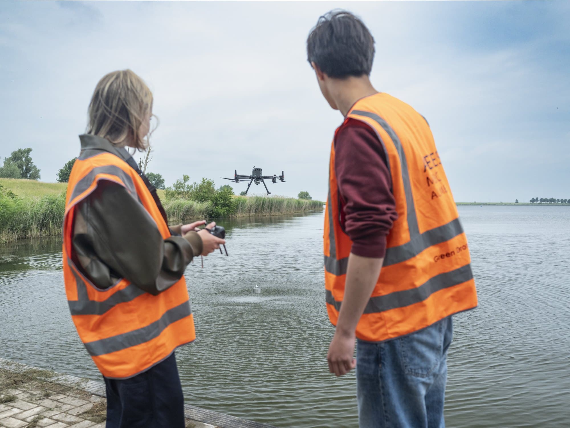 Twee studenten in veiligheidshesje vliegen een drone met een meetsonde boven het water, de sonde hangt net boven het wateroppervlak