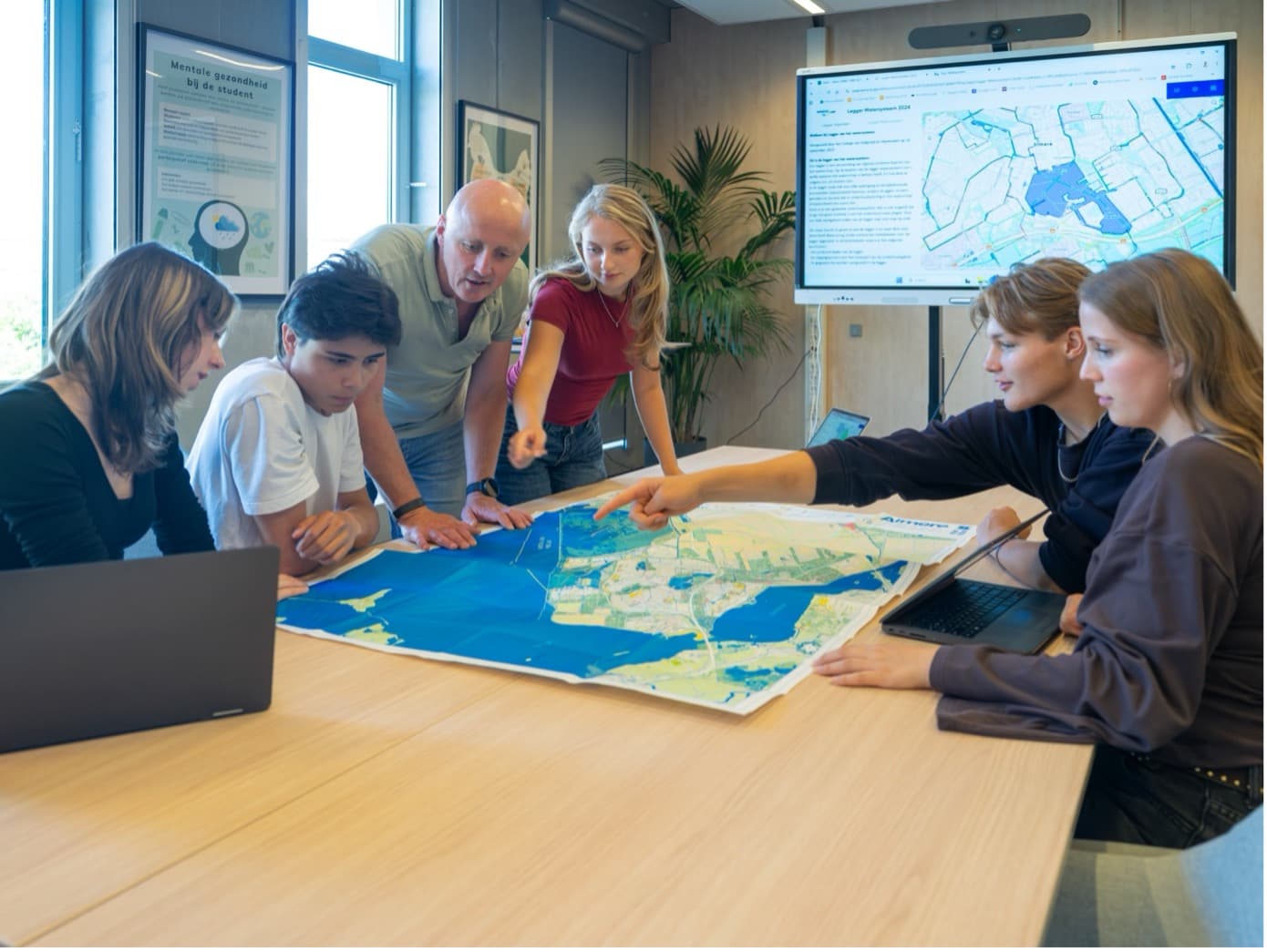 Studenten, een docent en medewerkers buigen zich samen over een grote waterkaart van Almere, met een digitaal scherm op de achtergrond