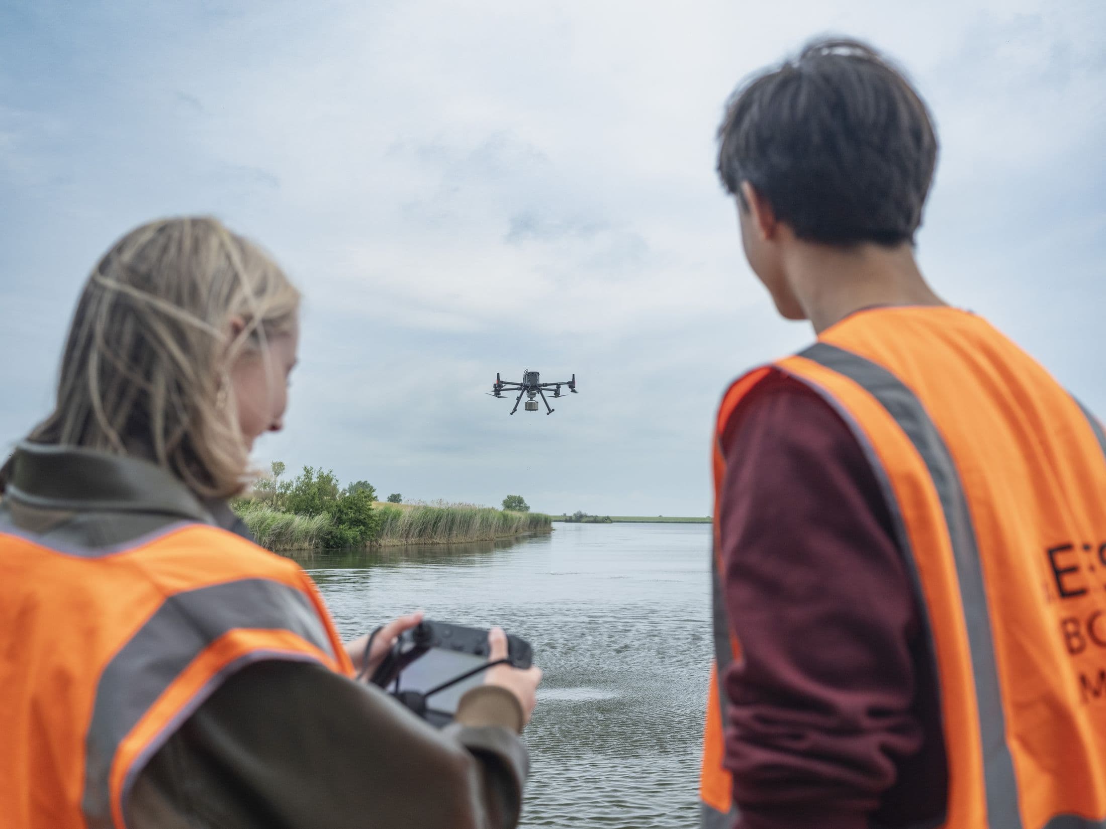Twee studenten in veiligheidshesje vliegen een drone boven het water